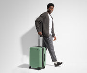 A 6'1" male model is standing next to and walking with The Bigger Carry-On suitcase by holding the raised handle to show size and scale