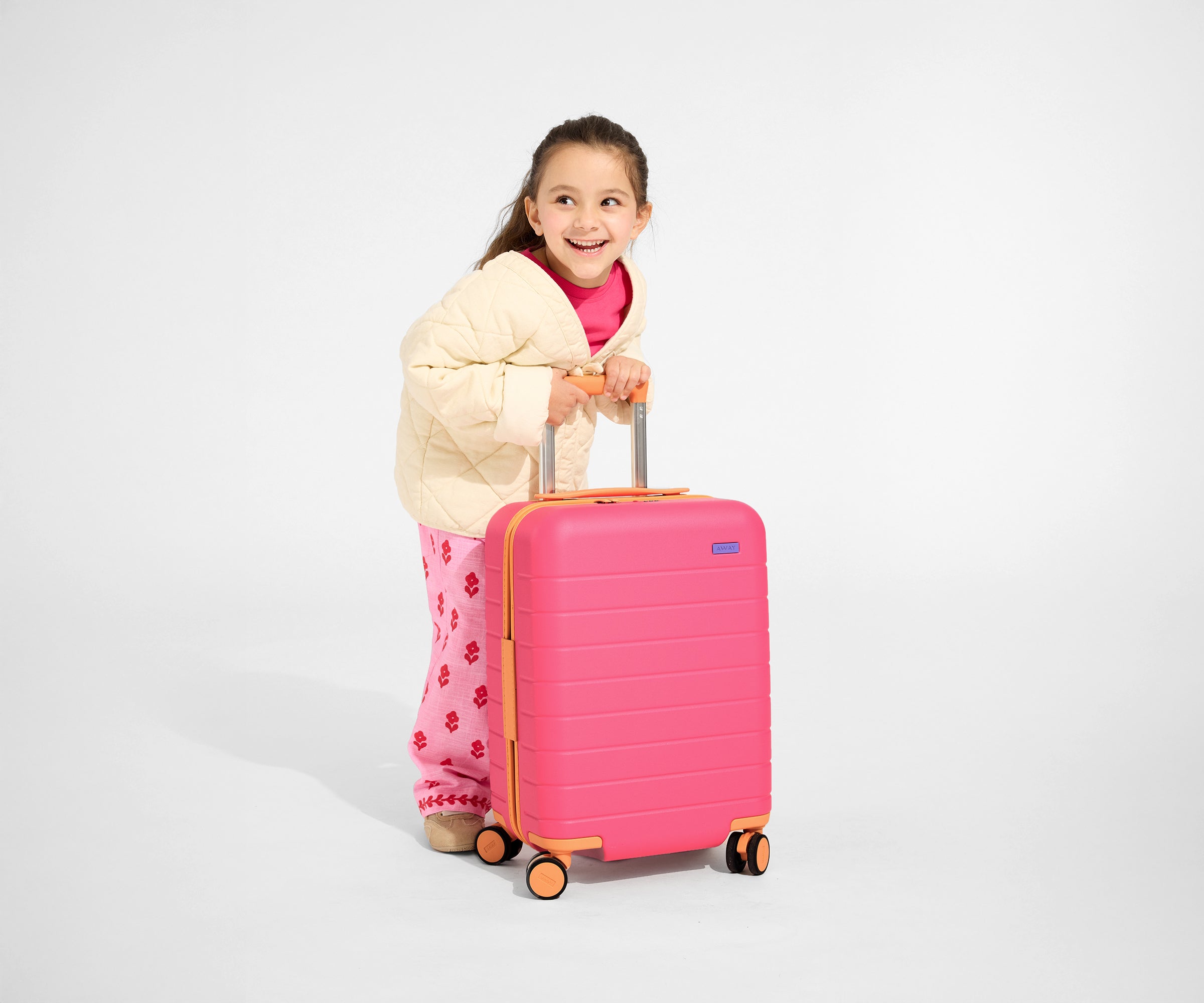 A child model is leaning on the raised handle of The Kids Carry-On suitcase in Cotton Candy Pink to show size and scale