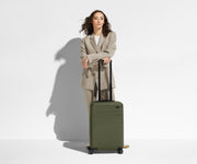 A 5'6" female model is standing next to The Bigger Carry-On suitcase and leaning her arms on the raised handle to show size and scale