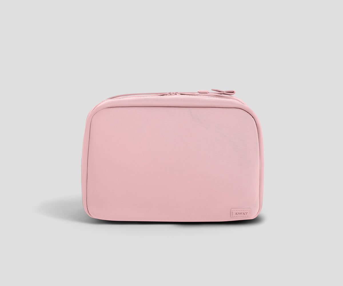 A light pink zippered toiletry bag with rounded corners and a small rectangular "Away" brand tag on the bottom right, shown against a solid light gray background.