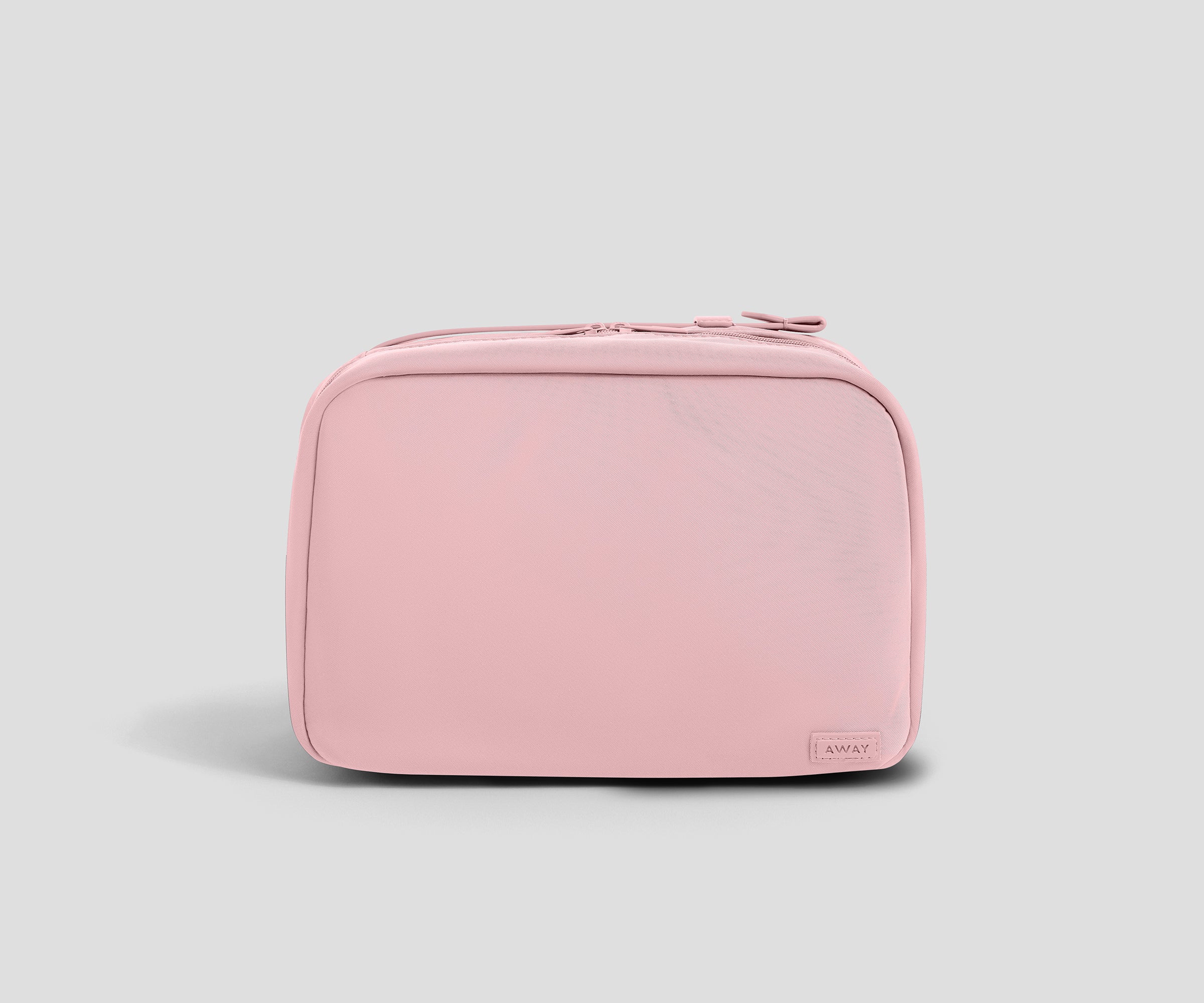 A light pink zippered toiletry bag with rounded corners and a small rectangular "Away" brand tag on the bottom right, shown against a solid light gray background.
