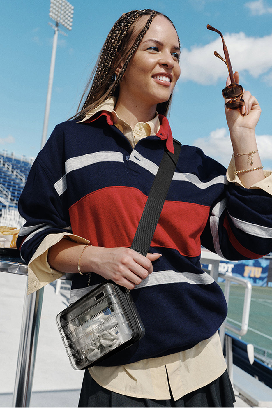 Woman in a colorful striped sweater holding sunglasses and a bag, with stadium seats in the background.